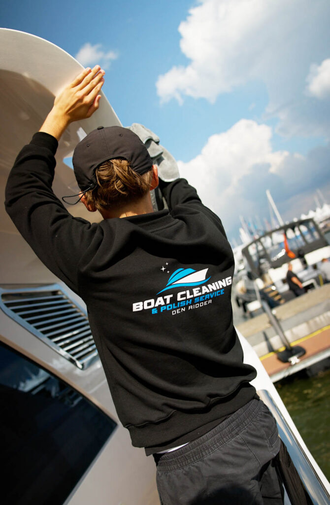 Persoon in zwarte cap en kleding houdt wit surf-/paneel omhoog op een boot, haven op achtergrond.